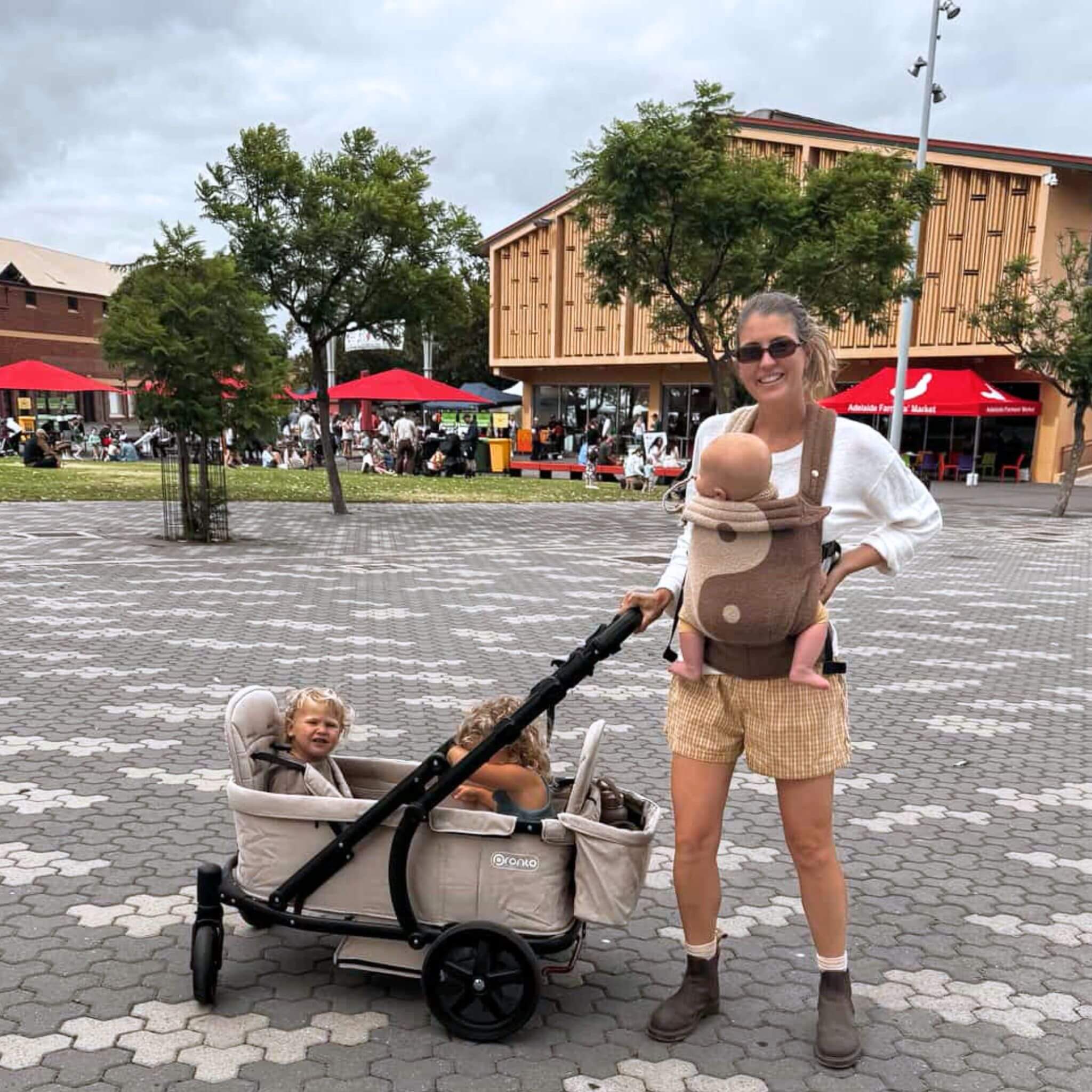 A woman pushing a beige Pronto Stroller with two children inside while carrying a baby in a carrier.